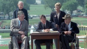 President George H.W. Bush signs the Americans with Disabilities Act into law, surrounded by two wheelchair users and two people standing behind him.