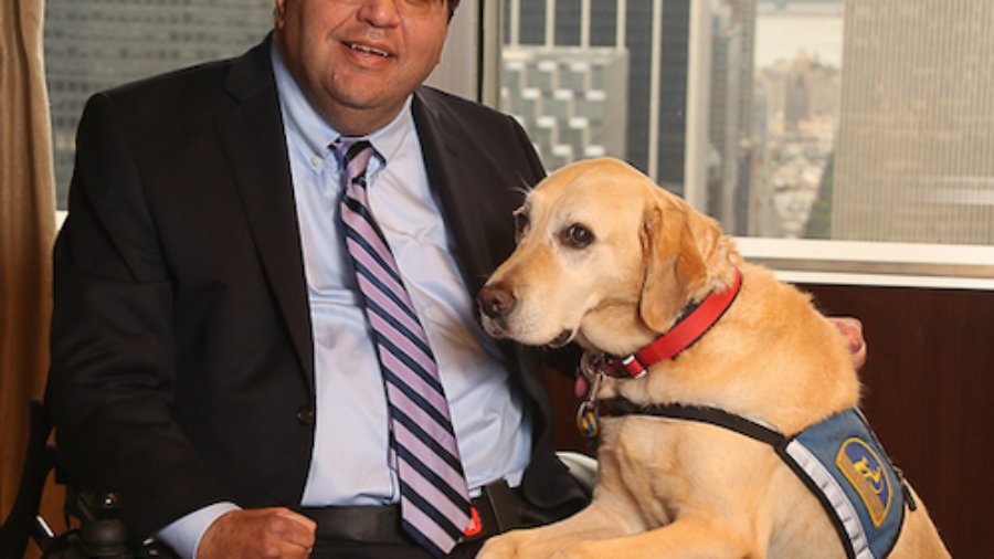 Jim Sinocchi sitting with a service dog in front of a window with skyscrapers behind him.