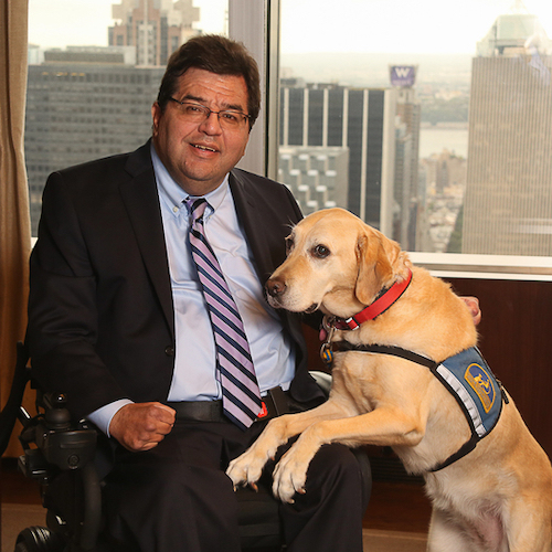 Jim Sinocchi sitting with a service dog in front of a window with skyscrapers behind him.