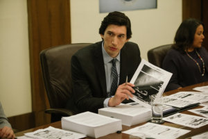 Adam Driver holding a paper with a photo on it, sitting at a desk with lots of papers on it