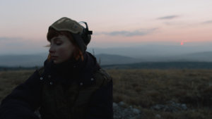 A woman with goggles on her head stands in the middle of an empty field