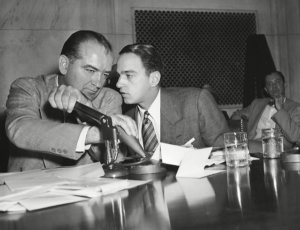 Roy Cohn and another man having a conversation at a desk with lots of papers and a hole puncher on it.