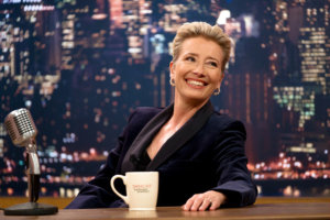 Emma Thompson sits behind a late night talk show desk with a city skyline background and a mug in front of her