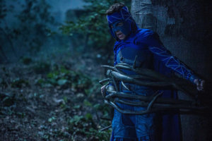 Mihajlo Milavic as Shade in a blue superhero outfit tied to a tree in the woods