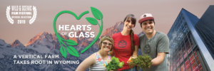 Three employees at Vertical Harvest holding plants in front of the greenhouse and mountains. Text: Hearts of Glass A Vertical Farm Takes Root in Wyoming