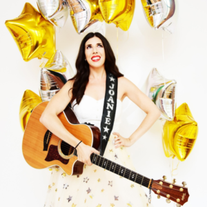 Joanie Leeds holding a guitar with the strap saying Joanie on it in front of yellow star balloons