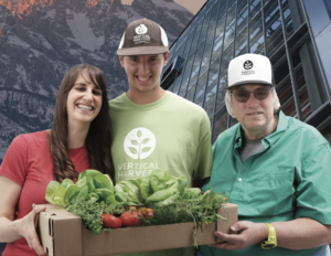 Three stars of Hearts of Glass smiling together in front of mountains and a greenhouse holding plants.