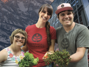 Three stars of Hearts of Glass smiling together in front of mountains and a greenhouse holding plants.