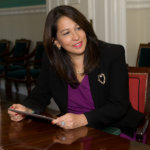 Carol Robles-Román sitting at a table holding an iPad, smiling looking to her left