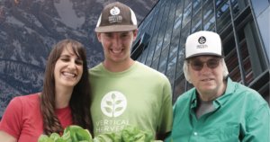 Three stars of Hearts of Glass smiling together in front of mountains and a greenhouse holding plants.