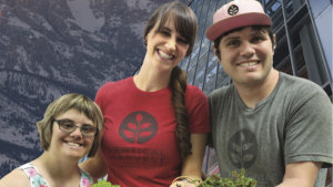 Three stars of Hearts of Glass smiling together in front of mountains and a greenhouse holding plants.