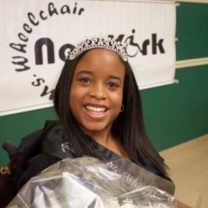 Katrina Hazell wearing a crown in front of a banner for Miss Wheelchair New York