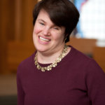 Rabbi Lauren Tuchman wearing a purple shirt inside a synagogue with the background blurred.