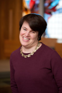 Rabbi Lauren Tuchman wearing a purple shirt inside a synagogue with the background blurred.