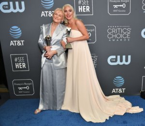 Glenn Close and Lady Gaga holding their Critics' Choice Awards, smiling in front of a banner with logos for the awards, AT&T and the CW.