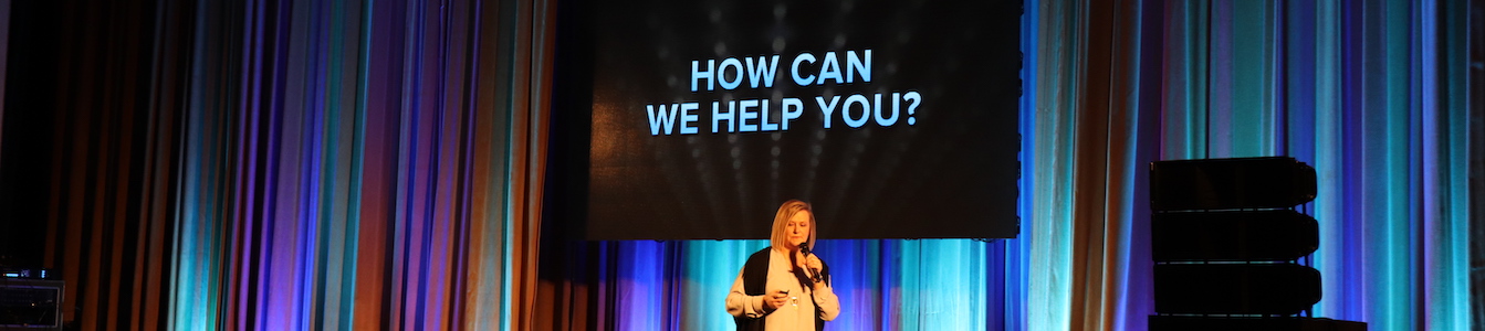 Jackie Lau giving a presentation on stage. the screen behind her says "How Can We Help You". Credit for image: Jackie Lau