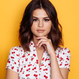 Selena Gomez wearing a white blouse with a cherry print posing for the camera with a yellow background