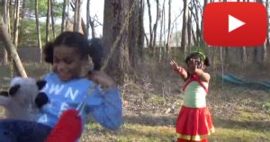 Still from Saylor and Selena with the two girls playing in a park, one on a swing being pushed by the other. YouTube play icon in upper right