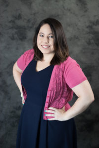 Stacey Herman smiling with her hands on her hips in front of a grey wall