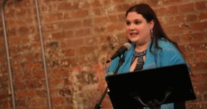 Rachel Kunstadt behind a microphone and a music stand and in front of a brick wall