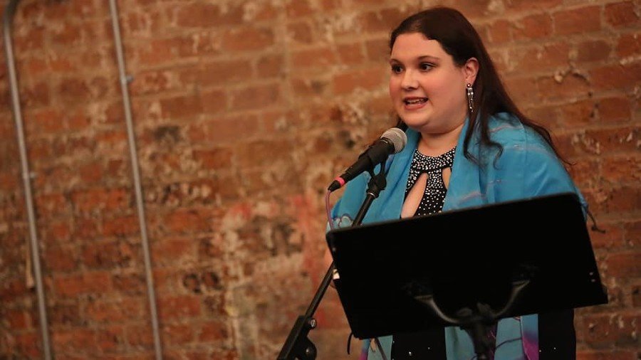 Rachel Kunstadt behind a microphone and a music stand and in front of a brick wall