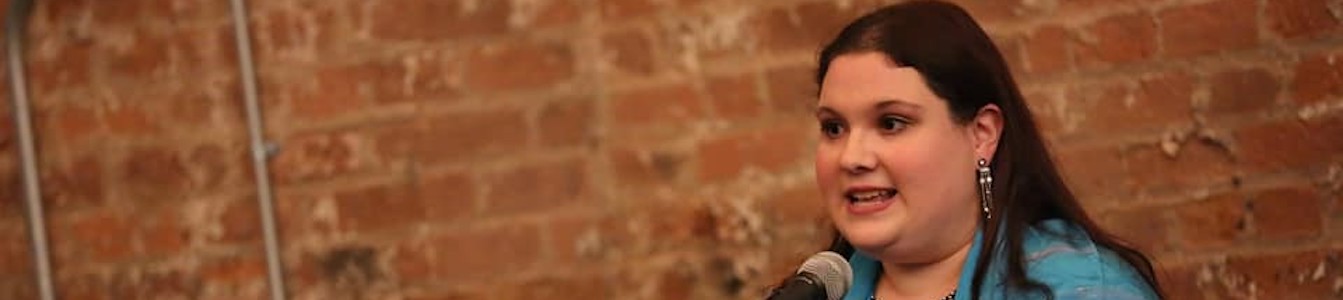 Rachel Kunstadt behind a microphone and a music stand and in front of a brick wall