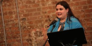 Rachel Kunstadt behind a microphone and a music stand and in front of a brick wall