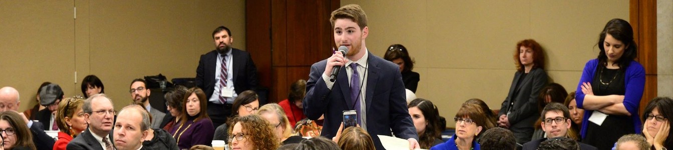 Adam Fishbein speaking with a microphone and papers in his hands, standing, as a large group of other people surround him