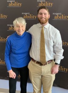 Eleanor Clift and Adam Fishbein smile in front of the RespectAbility banner with their arms around each other's backs