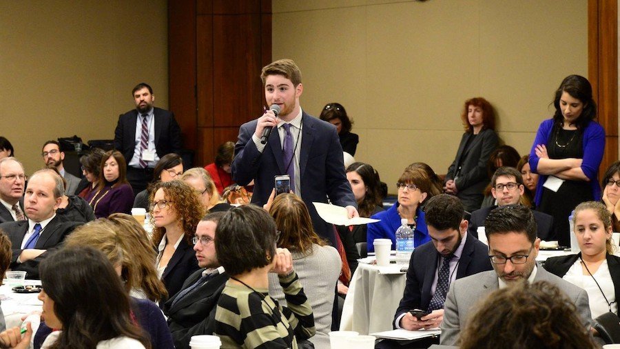 Adam Fishbein speaking with a microphone and papers in his hands, standing, as a large group of other people surround him