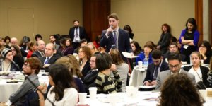 Adam Fishbein speaking with a microphone and papers in his hands, standing, as a large group of other people surround him