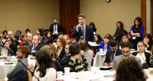 Adam Fishbein speaking with a microphone and papers in his hands, standing, as a large group of other people surround him