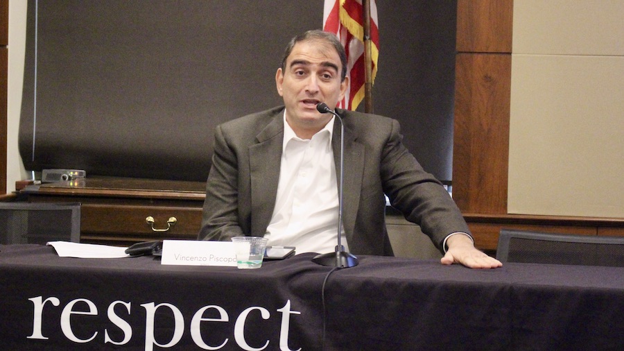 Vincenzo Piscopo speaking behind a microphone on a table with RespectAbility's logo on the tablecloth in front of a flag