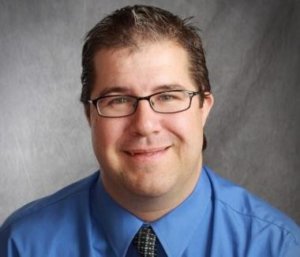 James Emmett smiling wearing a blue shirt and glasses in front of a grey backdrop