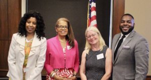 L-R: Janet LaBreck, Janie Jeffers, Diane Smith Howard, and Robert Stephens smiling in front of a wall and an American flag