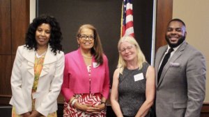 L-R: Janet LaBreck, Janie Jeffers, Diane Smith Howard, and Robert Stephens smiling in front of a wall and an American flag