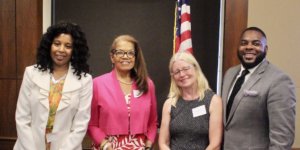 L-R: Janet LaBreck, Janie Jeffers, Diane Smith Howard, and Robert Stephens smiling in front of a wall and an American flag