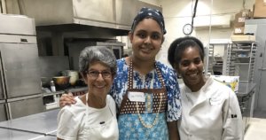 L-R: Sara Portman Milner, teen Sunflower program participant, Chef Marion Pitcher, smiling together with their arms around each other inside a kitchen