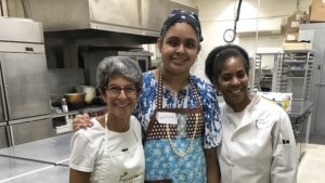 L-R: Sara Portman Milner, teen Sunflower program participant, Chef Marion Pitcher, smiling together with their arms around each other inside a kitchen