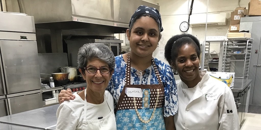 L-R: Sara Portman Milner, teen Sunflower program participant, Chef Marion Pitcher, smiling together with their arms around each other inside a kitchen