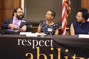 Matan Koch speaking as Laka Mitiku Negassa and Evelyn Kelly look on. All are sitting behind a table with RespectAbility's logo on the tablecloth and in front of an American flag