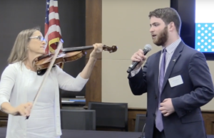 Adam Fishbein singing the national anthem in front of an American flag with Debbie Fink accompanying him on violin.