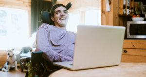 A young man who uses a wheelchair sits behind a laptop with the rest of his apartment blurred in the background