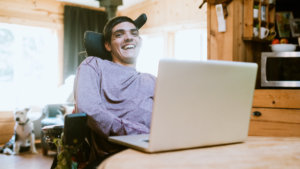 A young man who uses a wheelchair sits behind a laptop with the rest of his apartment blurred in the background