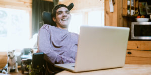 A young man who uses a wheelchair sits behind a laptop with the rest of his apartment blurred in the background