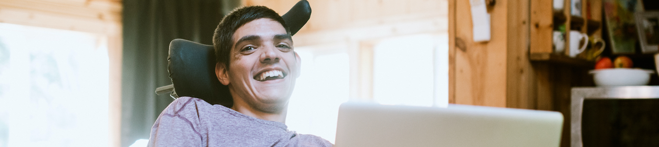 A young man who uses a wheelchair sits behind a laptop with the rest of his apartment blurred in the background