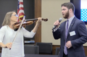Adam Fishbein singing the national anthem in front of an American flag with Debbie Fink accompanying him on violin.