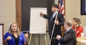 Ollie Cantos writing on a flip chart holding a walking stick, as Jennifer Laszlo Mizrahi and Steve Bartlett look on seated at a table. Sign language interpreter is in the lower left of the frame. American flag in the background