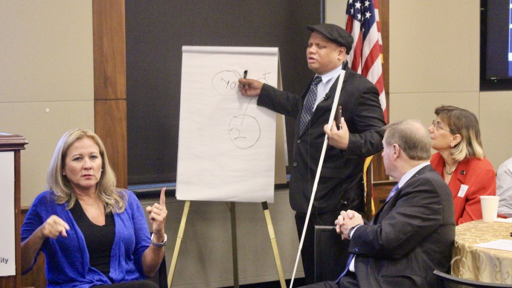 Ollie Cantos writing on a flip chart holding a large white cane, as Jennifer Laszlo Mizrahi and Steve Bartlett look on seated at a table. Sign language interpreter is in the lower left of the frame. American flag in the background