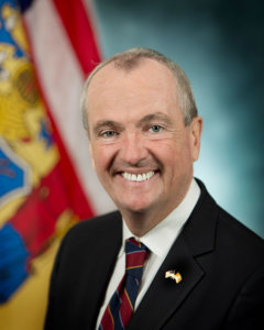 New Jersey Governor Phil Murphy smiling in front of the state flag.